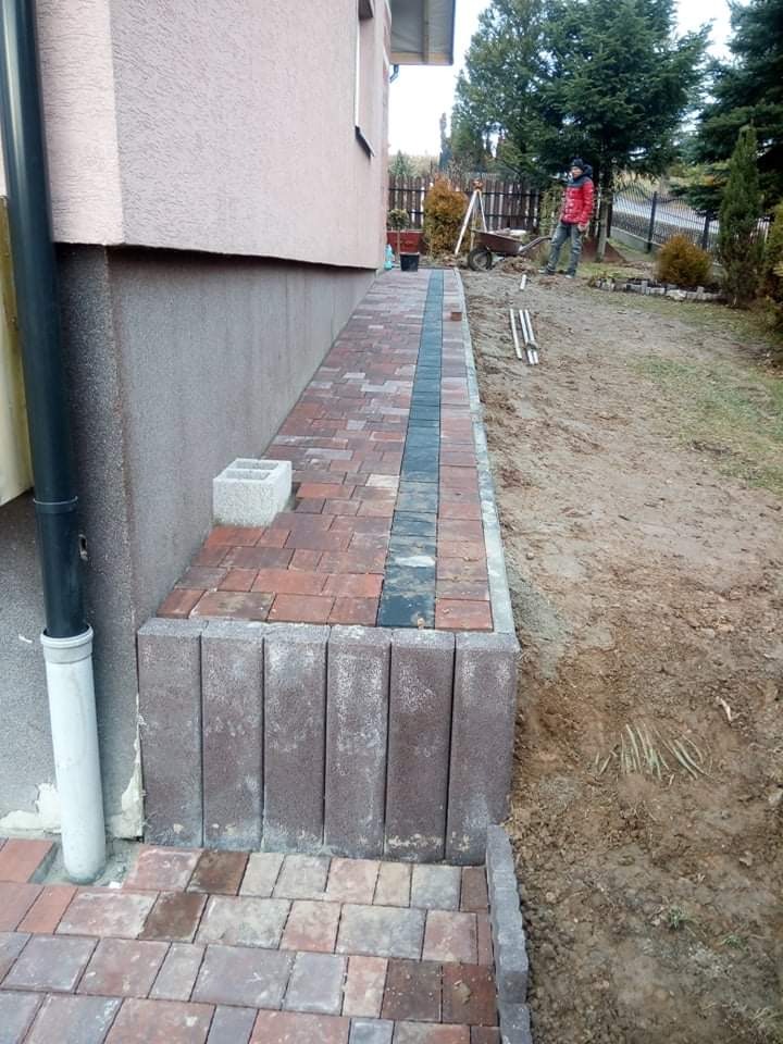 Fragment ścieżki z kostki brukowej wzdłuż ściany budynku, z widocznym obrzeżem i elementami czarnych kostek w centralnej części, w tle osoba z taczką.