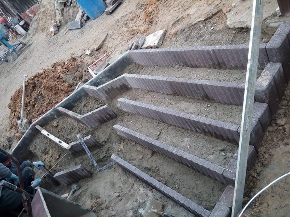 Budowa schodów z kostki brukowej na nierównym terenie, widoczne szare kostki, ziemia, poziomica i pracownik w rękawicach.