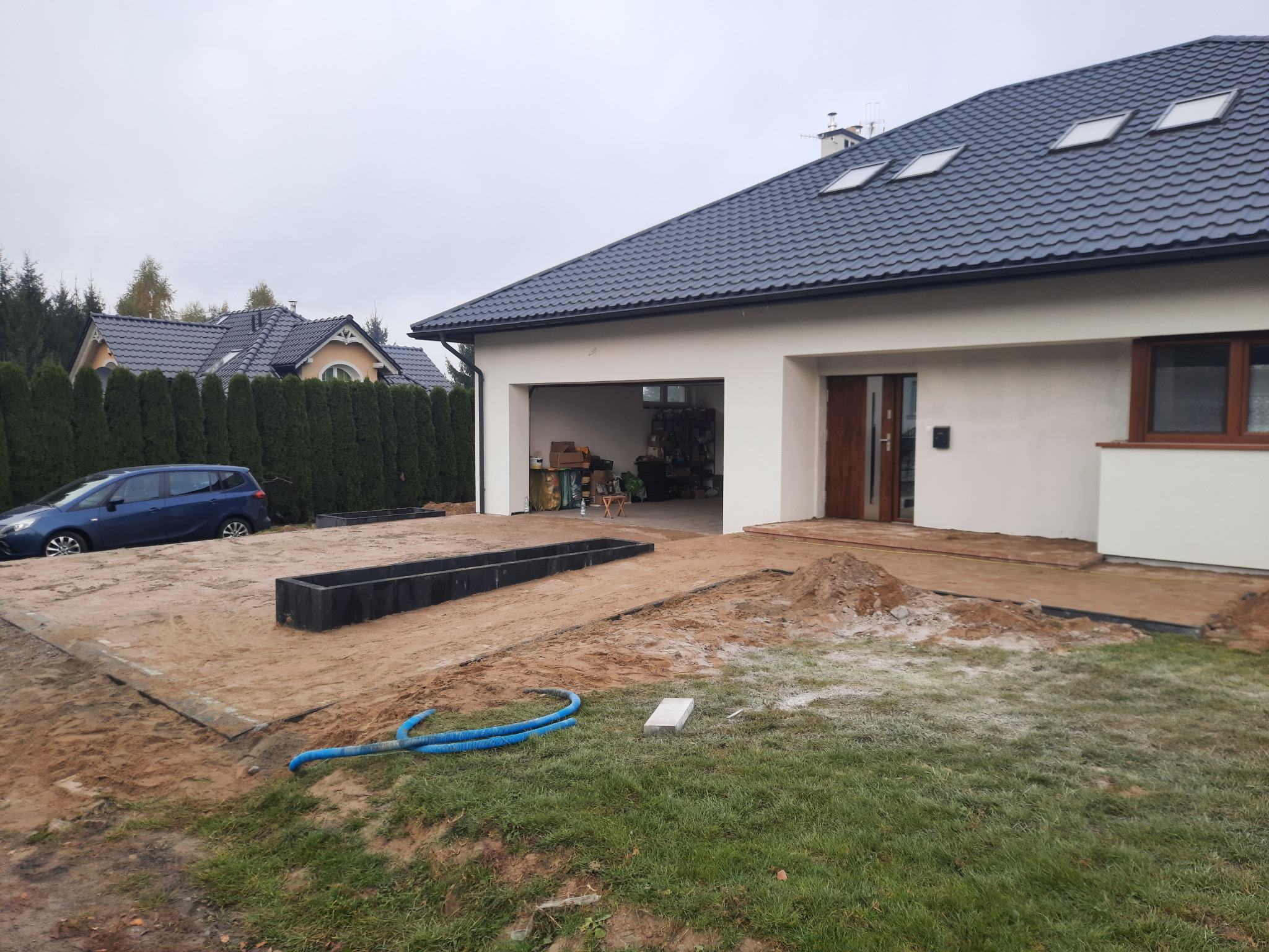Przygotowanie podłoża pod kostkę brukową przed nowoczesnym domem z garażem, widoczne czarne obrzeża i nawodniony piasek, błękitny wąż ogrodowy na trawie.