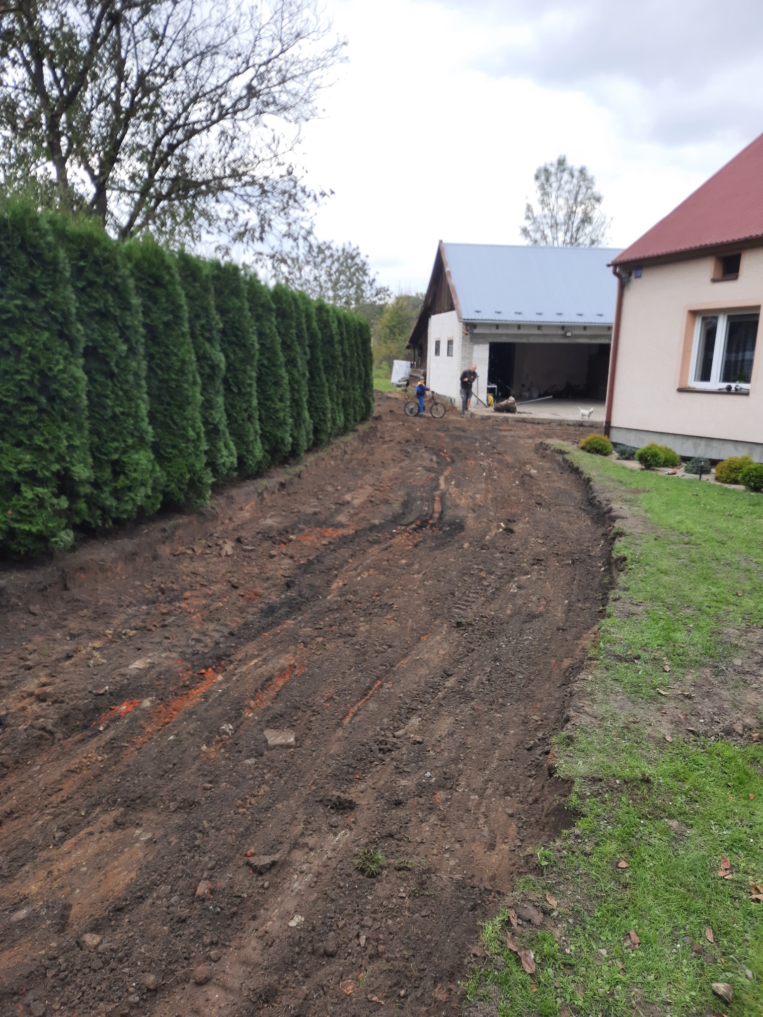 Przygotowany teren pod budowę drogi dojazdowej, widoczne ślady opon, wyrównana ziemia, w tle żywopłot z tui, budynki gospodarcze i mieszkalne, pochmurne niebo.