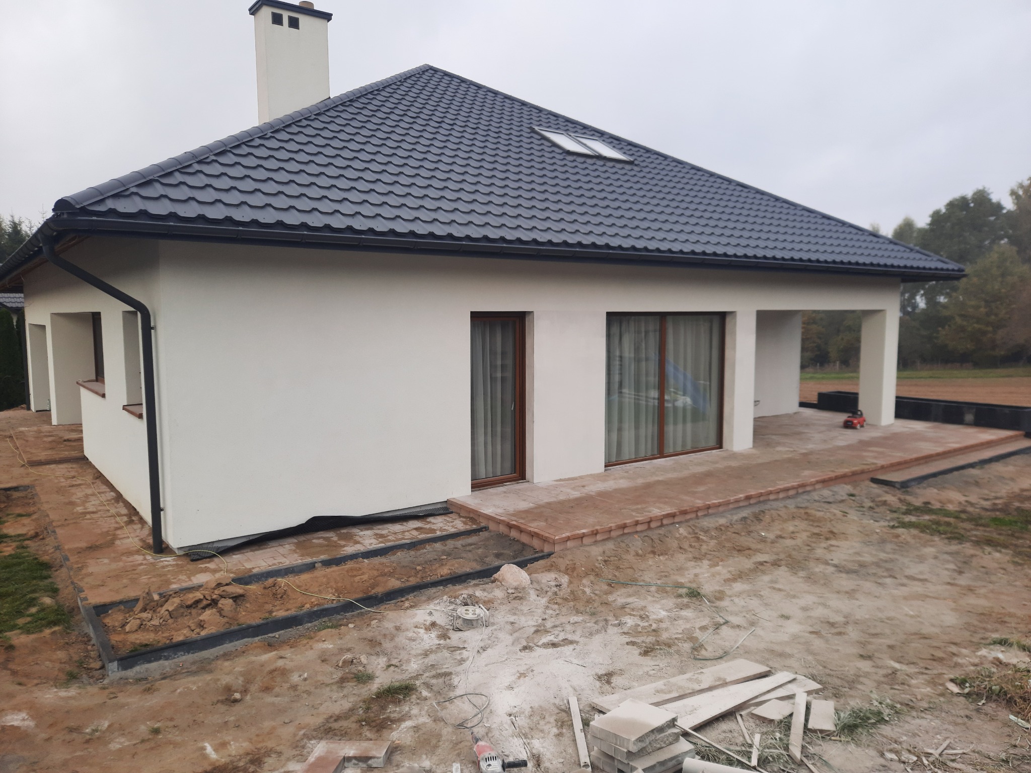 Nowoczesny dom jednorodzinny z ciemną dachówką, częściowo wyłożonym tarasem z ceglanej kostki, widoczne przygotowania do dalszego układania kostki brukowej na posesji.