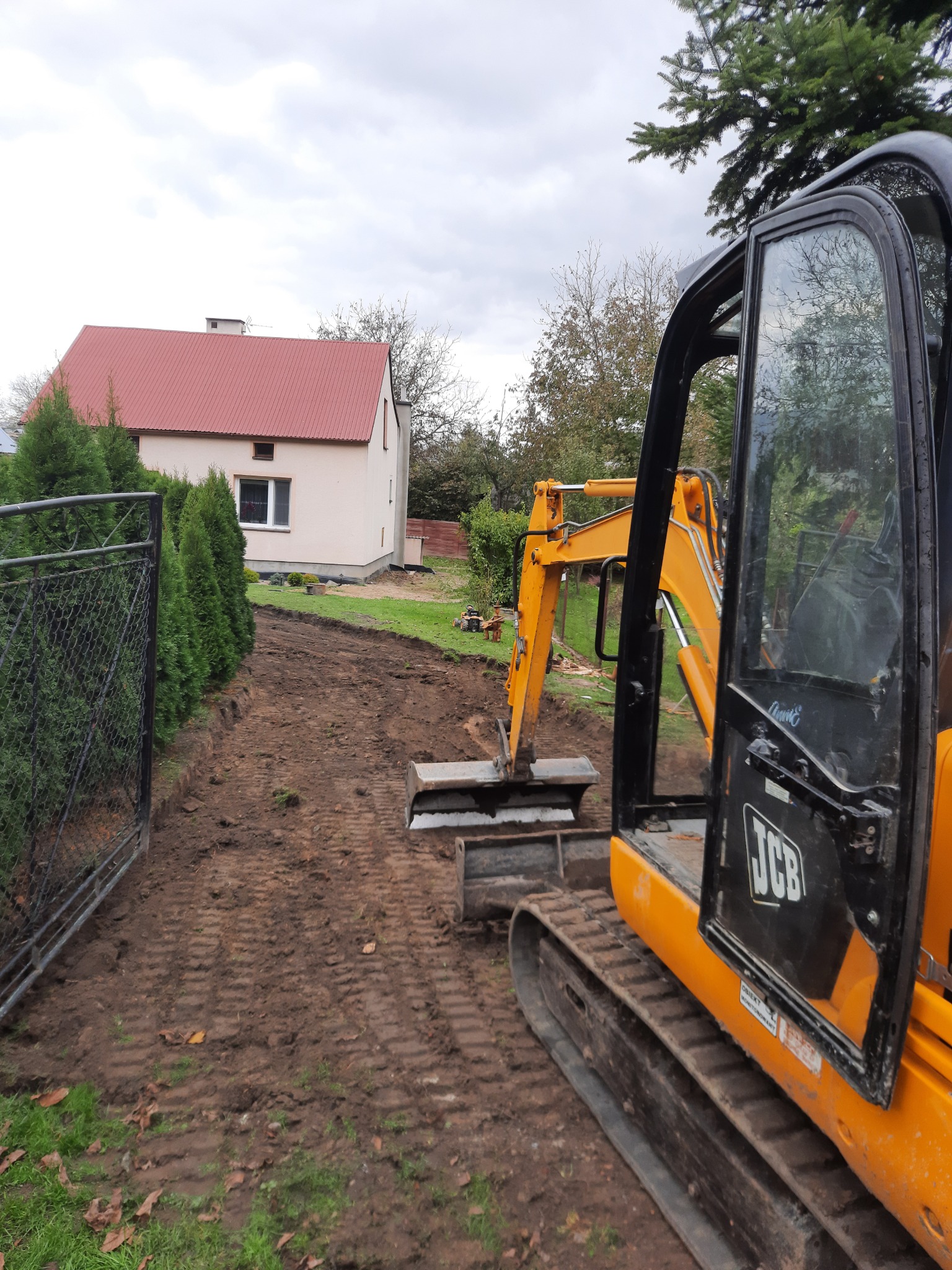 Żółta minikoparka JCB z otwartą kabiną na działce z wykopanym pasem ziemi, widoczny dom z czerwoną dachówką i rząd tuj przy ogrodzeniu.