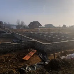 Wykop pod fundamenty z wymurowanymi ścianami z bloczków betonowych, widoczne wystające pręty zbrojeniowe, deska i folia budowlana na ziemi, w tle zabudowa jednorodzinna i poranna mgła.