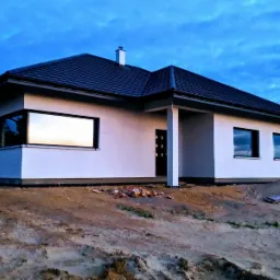 Niedawno ukończony, parterowy dom murowany z ciemną dachówką, białymi ścianami i dużymi oknami, widoczne przygotowania terenu wokół budynku, w tle las i błękitne niebo.