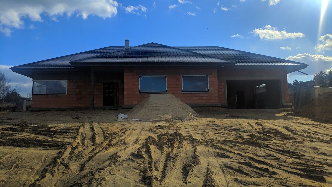Niedokończony dom jednorodzinny z czerwonej cegły, z ciemnym dachem, z oknami wstawionymi w surowy mur, z kupką piasku przed budynkiem i widocznymi śladami opon na piaszczystym gruncie, w słoneczny...