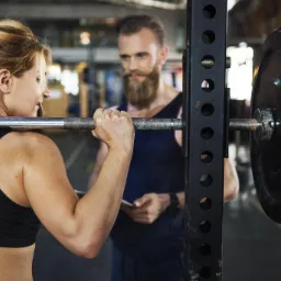 Kobieta przygotowuje się do podniesienia sztangi pod okiem brodatego trenera z notesem w dłoniach.