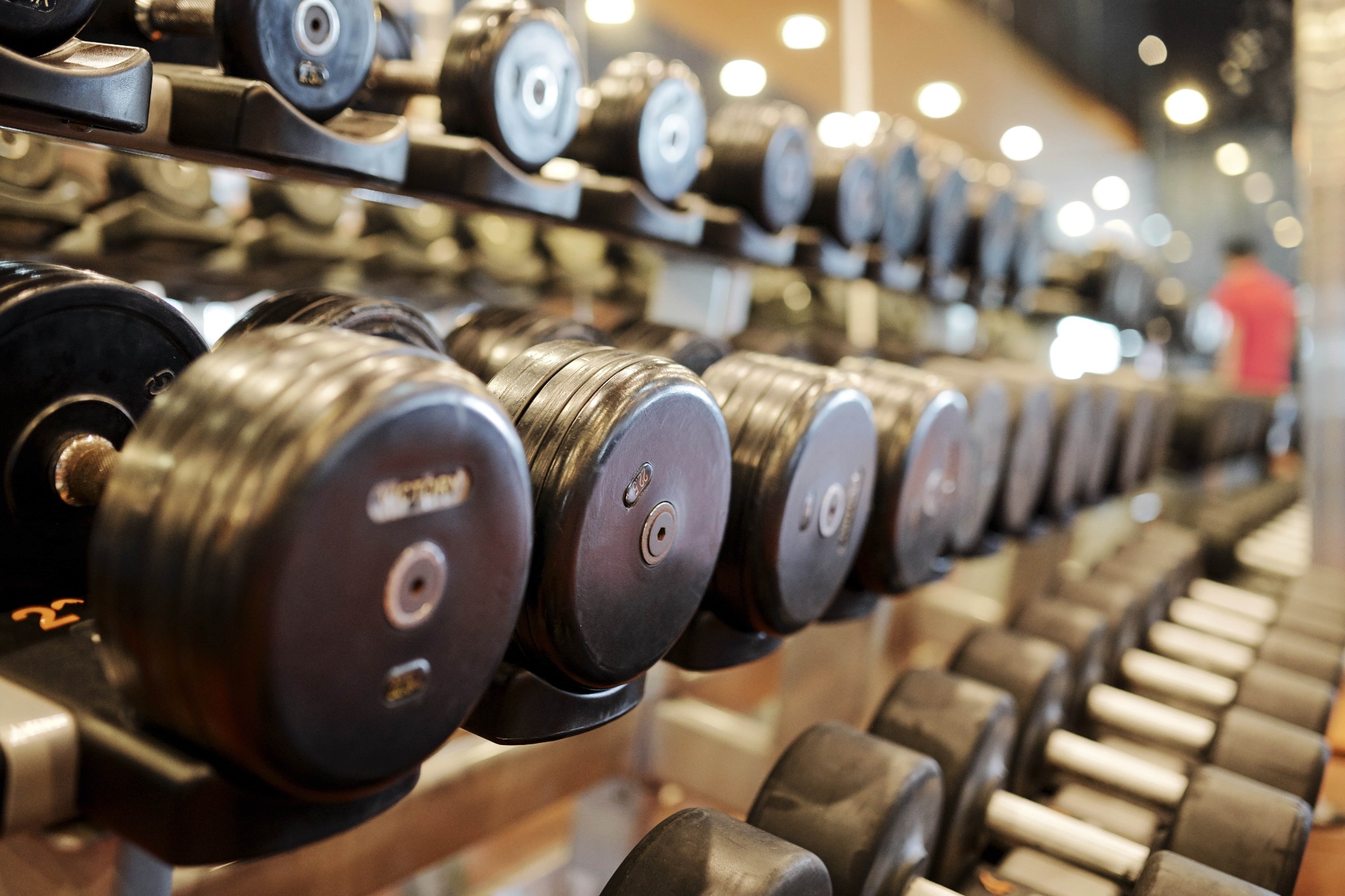 Rząd hantli na stojaku w siłowni, z widocznymi oznaczeniami wagowymi i elementami mocującymi, w tle rozmyta sylwetka osoby.
