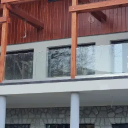 Szklana balustrada na balkonie domu z drewnianą konstrukcją dachu i elewacją z kamienia naturalnego.