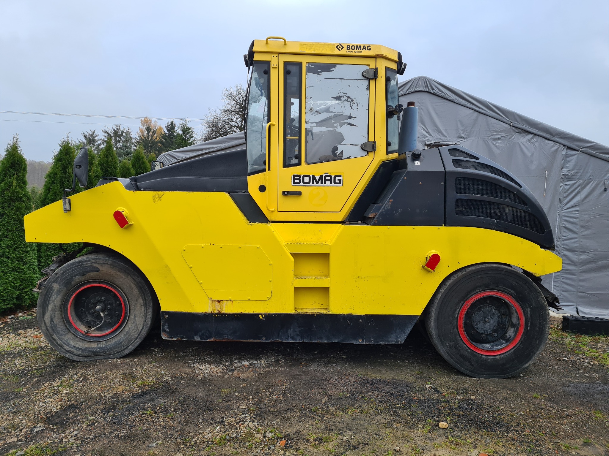 Żółto-czarny walec drogowy marki Bomag, widok z boku, z widocznymi oponami z czerwonymi felgami, stojący na żwirowym podłożu, z zarysowaniami na szybie kabiny i plandeką w tle.