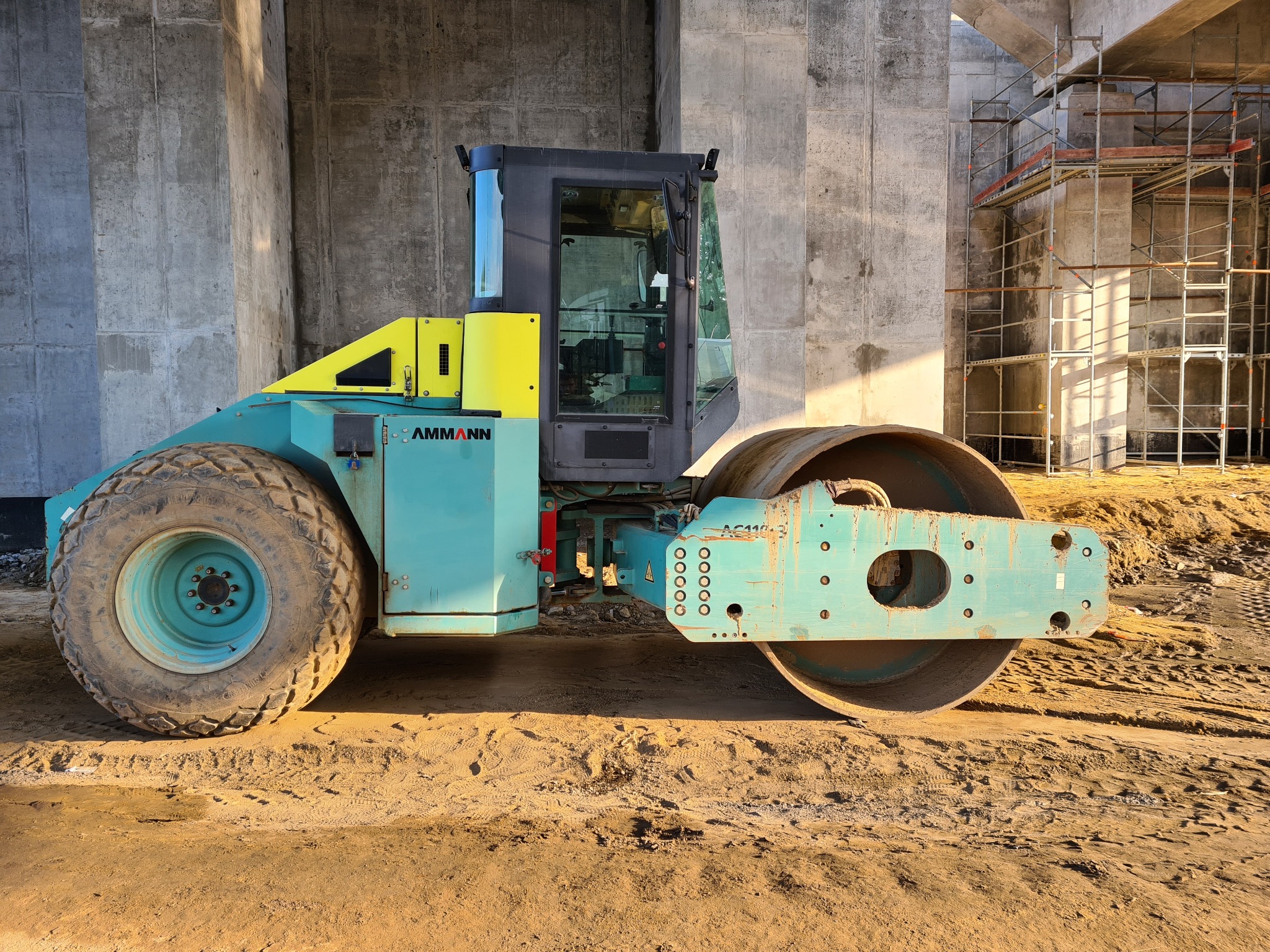 Niebiesko-żółty walec drogowy Ammann AC110 na piaszczystym gruncie pod betonowym wiaduktem w budowie, w tle widoczne rusztowania.
