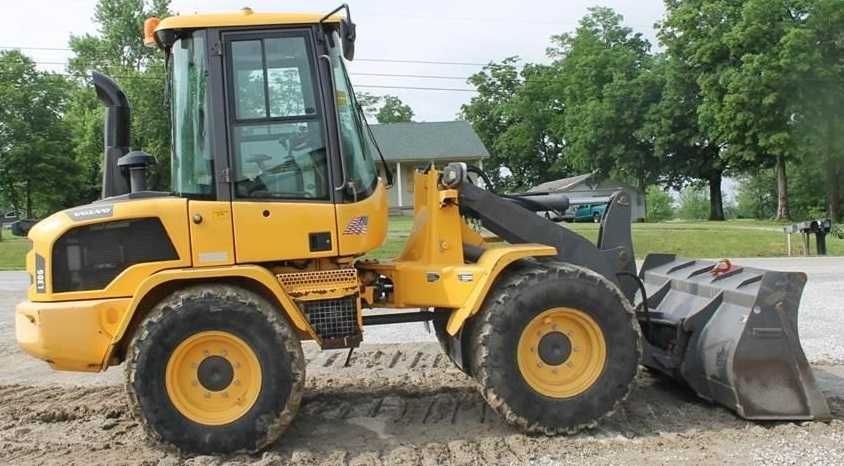 Żółta ładowarka kołowa Volvo L30G stoi na żwirowym podłożu, z widoczną kabiną operatora i łyżką, na tle zieleni drzew i domu.