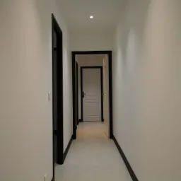 Długi, jasny korytarz z białymi ścianami, szarą podłogą z płytek i czarnymi futrynami drzwi, prowadzący do kolejnych pomieszczeń.