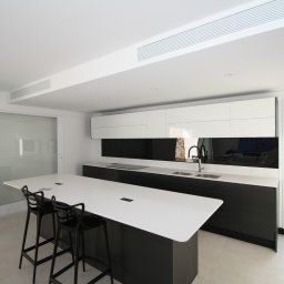 Krzysztof Nosek - Nowoczesna kuchnia w bieli i czerni z wyspą i wysokimi krzesłami barowymi, minimalistyczne szafki wiszące i czarny panel nad blatem, jasna podłoga i duże okno z widokiem na zieleń.