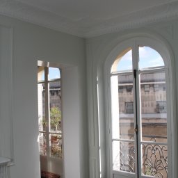 Krzysztof Nosek - Wnętrze pokoju z ciemną parkietową podłogą ułożoną w jodełkę, białymi ścianami z dekoracyjnymi panelami i listwami przysufitowymi, oraz wysokimi oknami z widokiem na budynek naprzeciwko.