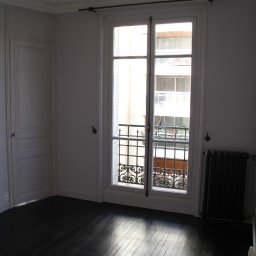 Krzysztof Nosek - Pusty pokój z ciemną, lakierowaną podłogą z desek, białymi ścianami z listwami przysufitowymi, oknem balkonowym z ozdobną kratą i grzejnikiem żeliwnym pod oknem.