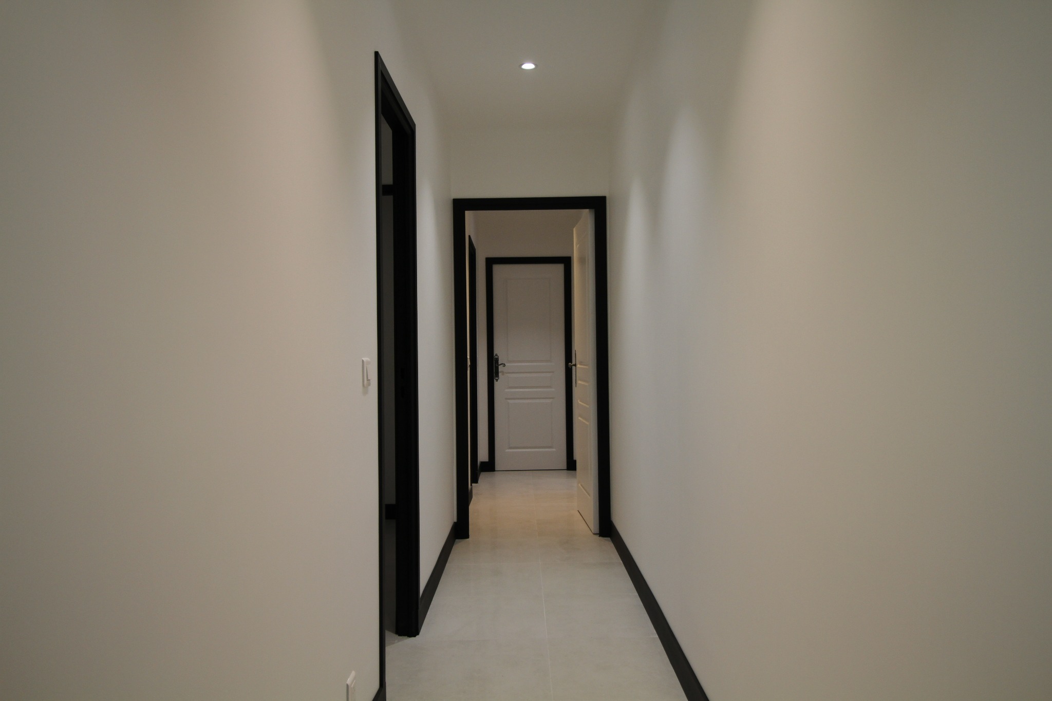 Długi, jasny korytarz z białymi ścianami, szarą podłogą z płytek i czarnymi futrynami drzwi, prowadzący do kolejnych pomieszczeń.