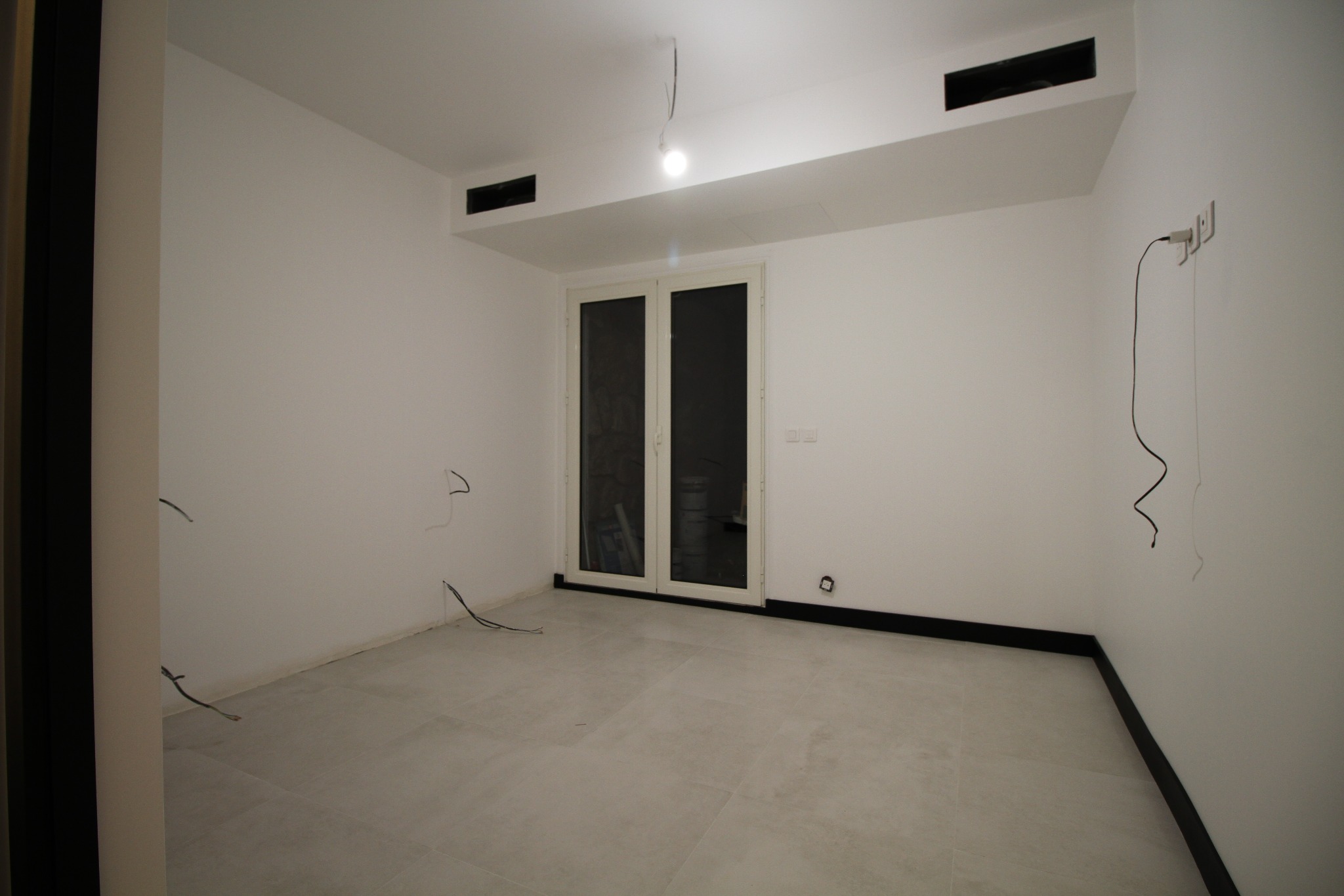 Pusty, jasny pokój z szarymi płytkami podłogowymi, czarną listwą przypodłogową i niedokończonymi przewodami elektrycznymi wystającymi ze ścian i sufitu; widoczne podwójne drzwi balkonowe i czarne...