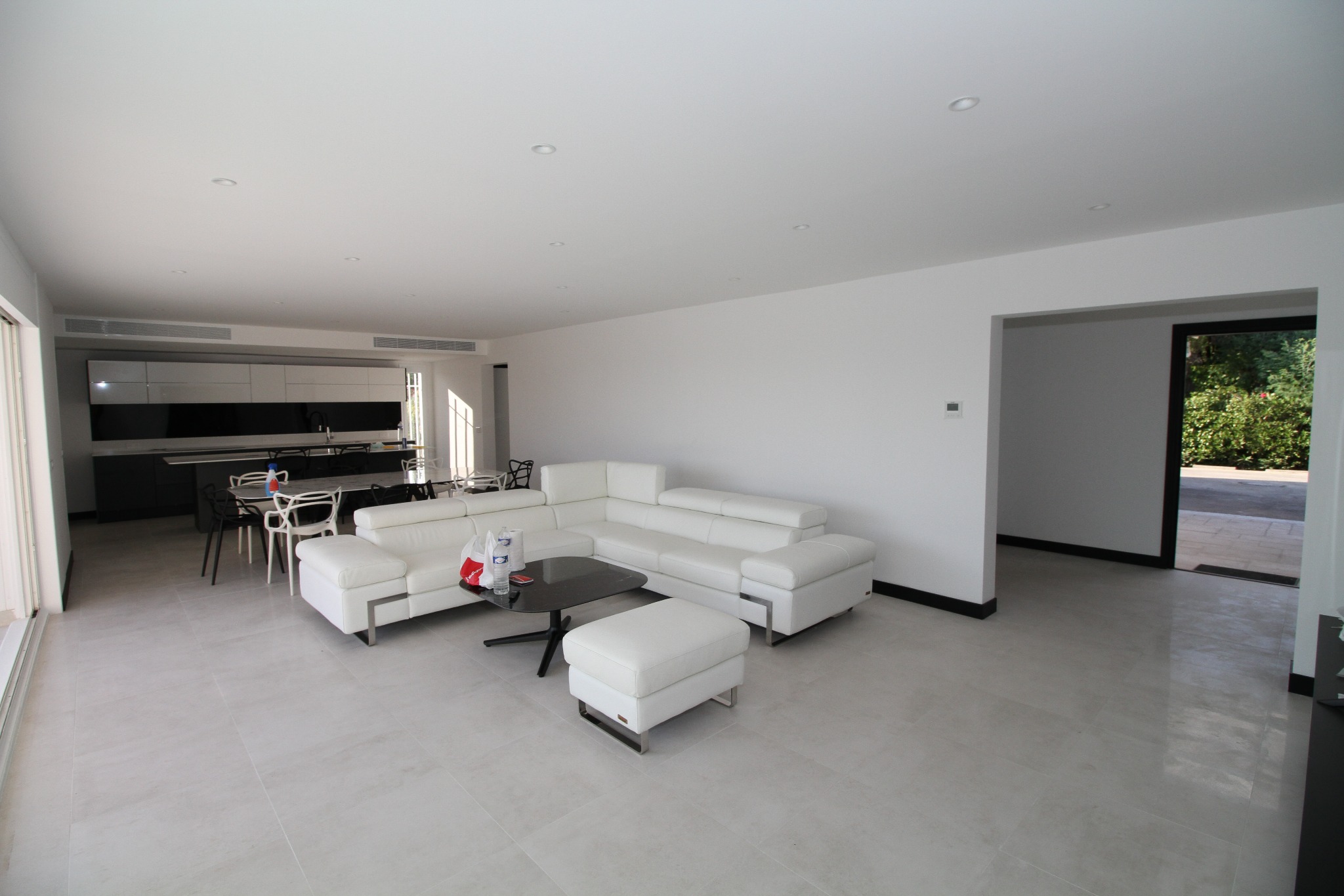 Przestronny salon z jasnymi, kwadratowymi płytkami podłogowymi, nowoczesną białą sofą narożną i czarnym stolikiem kawowym. W tle widoczna otwarta kuchnia z czarno-białymi szafkami.