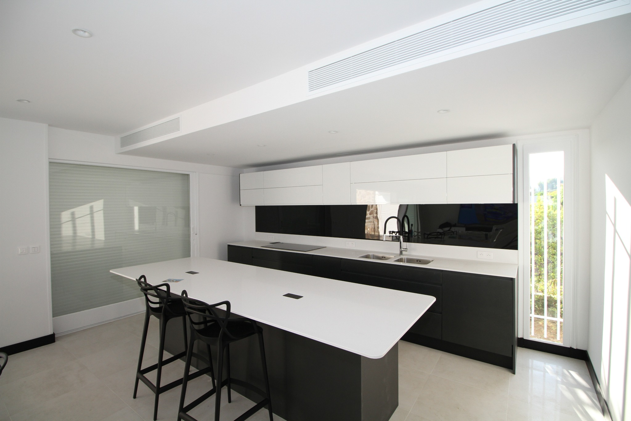 Nowoczesna kuchnia w bieli i czerni z wyspą i wysokimi krzesłami barowymi, minimalistyczne szafki wiszące i czarny panel nad blatem, jasna podłoga i duże okno z widokiem na zieleń.