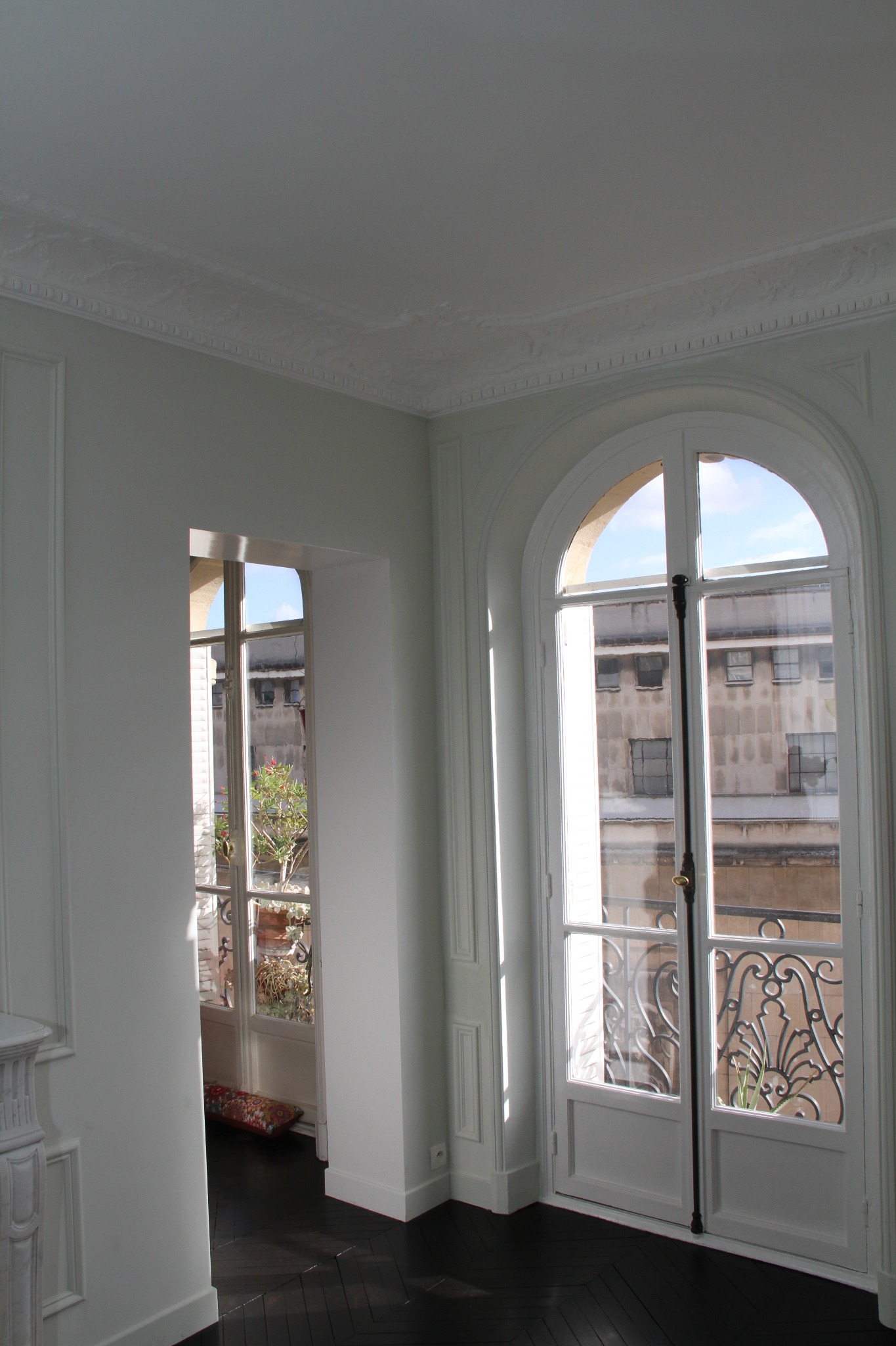 Wnętrze pokoju z ciemną parkietową podłogą ułożoną w jodełkę, białymi ścianami z dekoracyjnymi panelami i listwami przysufitowymi, oraz wysokimi oknami z widokiem na budynek naprzeciwko.