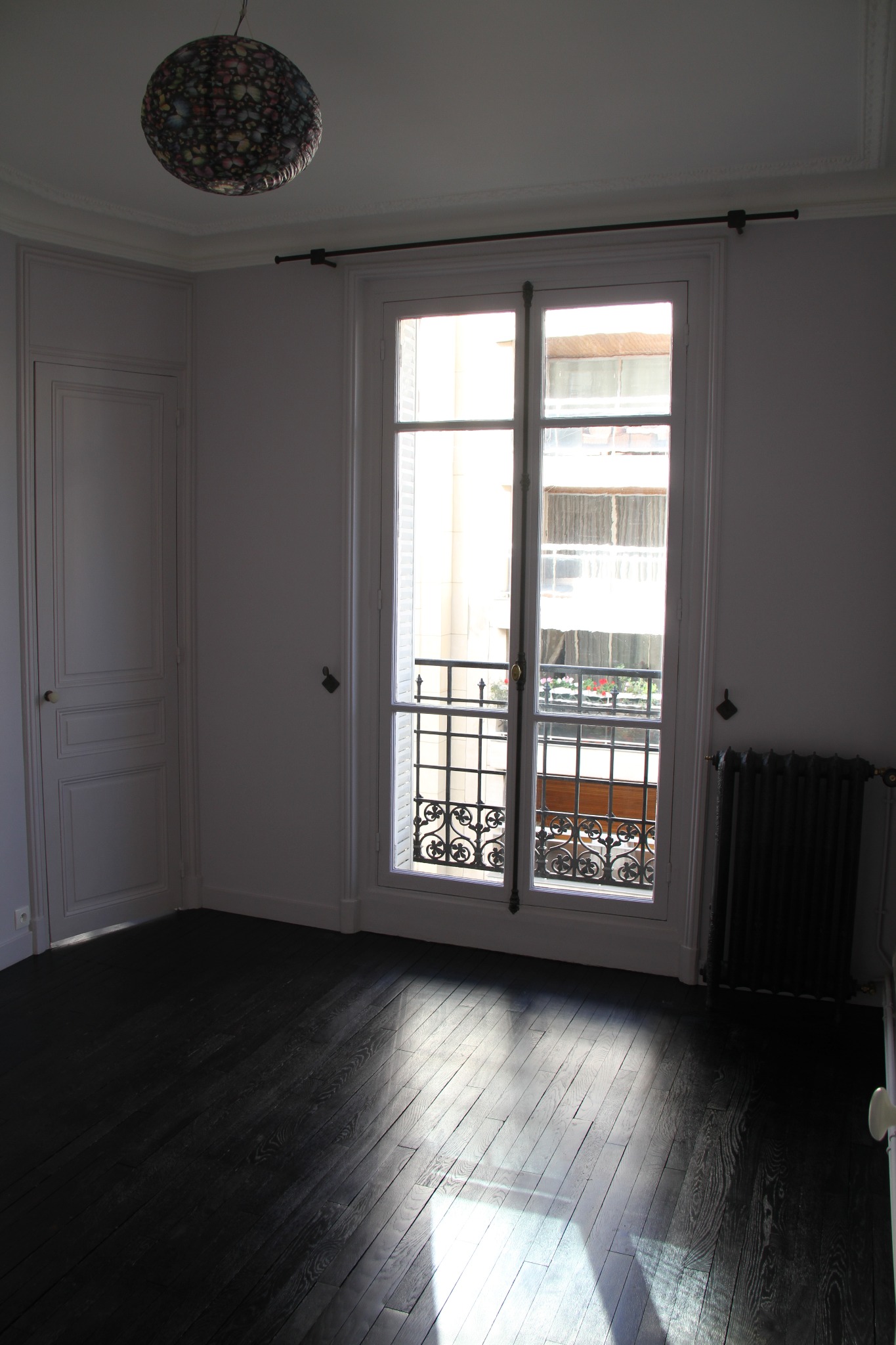 Pusty pokój z ciemną, lakierowaną podłogą z desek, białymi ścianami z listwami przysufitowymi, oknem balkonowym z ozdobną kratą i grzejnikiem żeliwnym pod oknem.