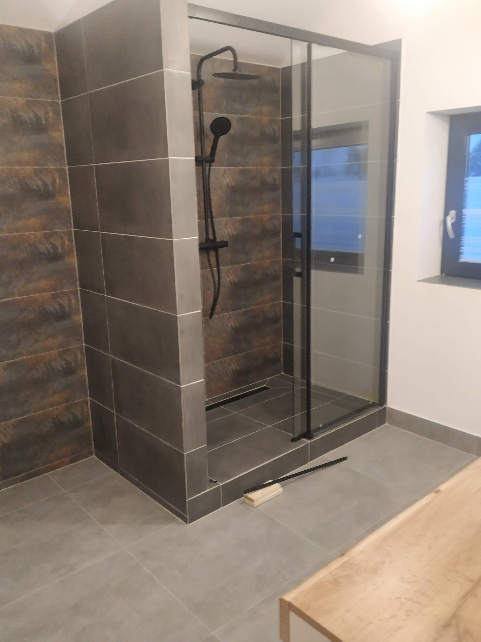 Nowoczesna kabina prysznicowa z ciemnymi płytkami imitującymi kamień, czarna armatura i profile, widoczny fragment podłogi i fragment blatu umywalki, na podłodze leżą narzędzia.