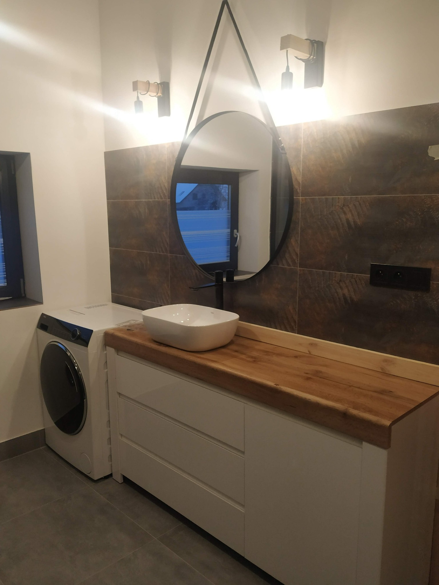 Nowoczesna łazienka z pralką obok szafki umywalkowej z blatem z litego drewna, owalne lustro na pasku, brązowe płytki imitujące kamień i minimalistyczne oświetlenie.