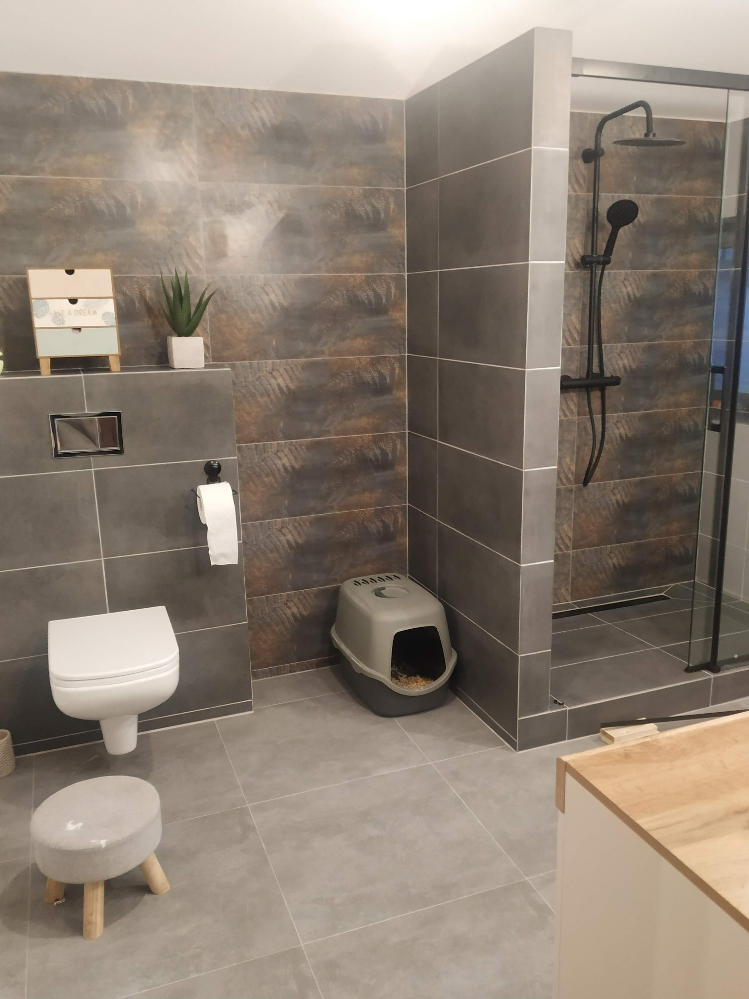 Wykończona łazienka z szarymi, poziomymi płytkami imitującymi wzór roślinny, kabiną prysznicową z czarną armaturą, toaletą, małym stołkiem i kuwetą dla kota.