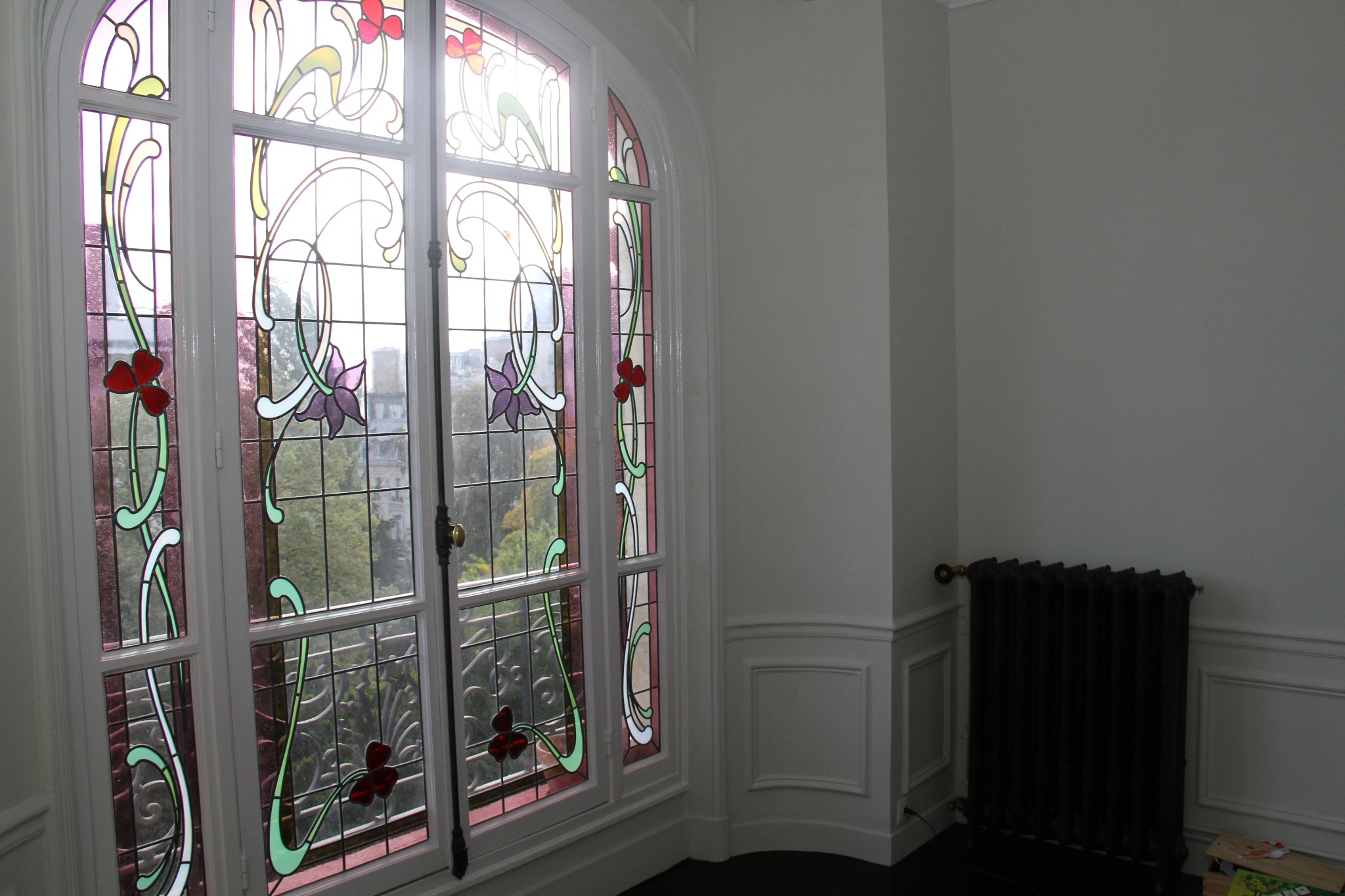 Okno z witrażami w stylu art nouveau, z motywami kwiatowymi w kolorach czerwonym i fioletowym, widok na zieleń za oknem, białe ramy okienne i ciemny kaloryfer w rogu pokoju.