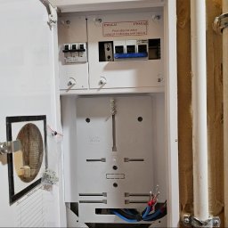 ELEKTROświatło - Otwarta biała rozdzielnica elektryczna ELMARO z widocznymi wyłącznikami nadprądowymi, przygotowanymi przewodami i ostrzeżeniem 'UWAGA!'. Obok biegną białe rury instalacyjne.