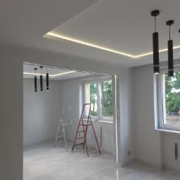 Jasne wnętrze w trakcie remontu z drewnopodobną podłogą, dwiema drabinami, oświetleniem LED w suficie podwieszanym i wiszącymi lampami tubowymi.