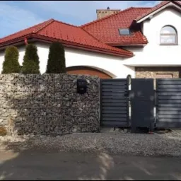 Nowoczesna posesja z ogrodzeniem gabionowym z kamienia, przesuwną bramą wjazdową z poziomych paneli i furtką w kolorze antracytowym, na tle domu z czerwoną dachówką i zielonymi tujami.