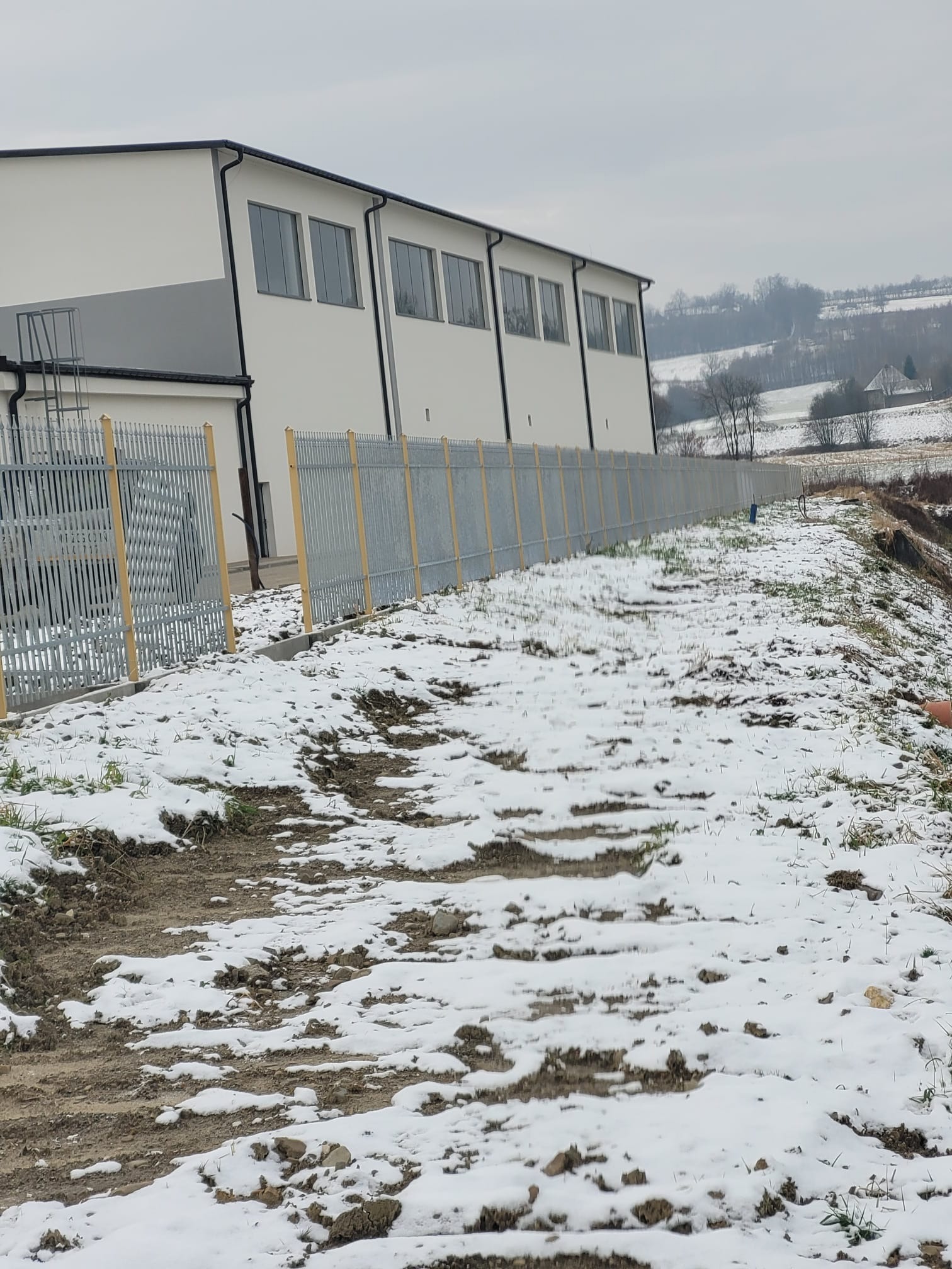 Długa, szara metalowa konstrukcja ogrodzeniowa z żółtymi słupkami, biegnąca wzdłuż budynku przemysłowego na tle zimowego krajobrazu z resztkami śniegu na ziemi.