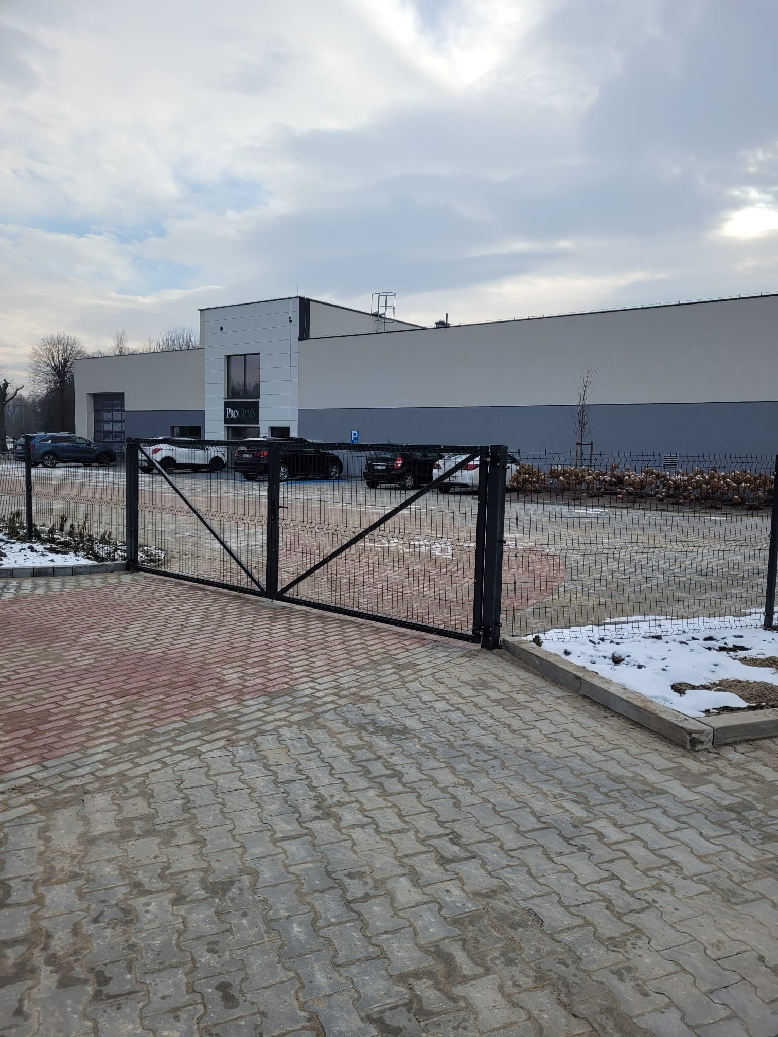 Otwarta, czarna brama wjazdowa z siatki ogrodzeniowej na tle budynku firmy z zaparkowanymi samochodami i resztkami śniegu.