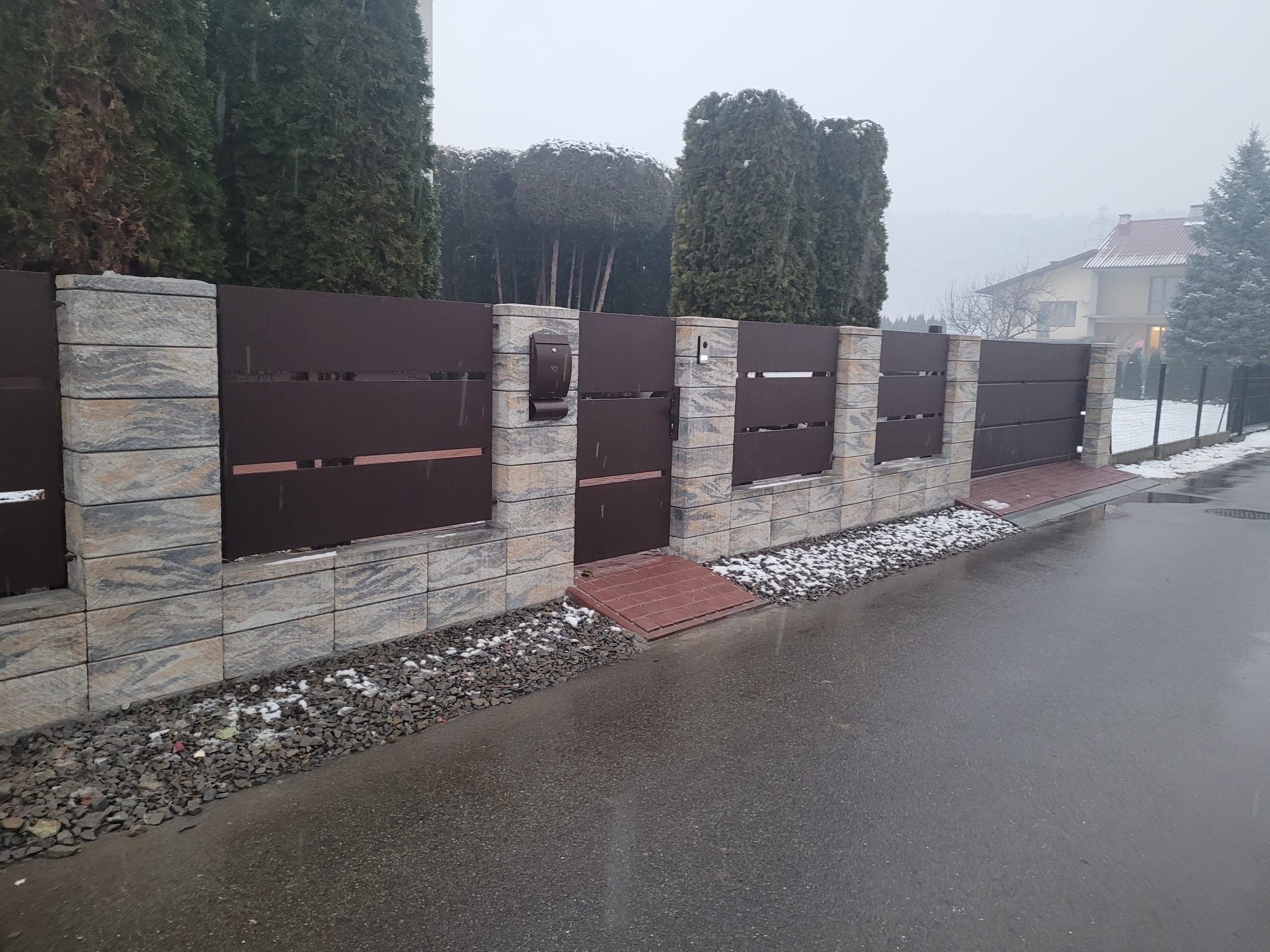 Nowoczesne ogrodzenie posesji z brązowymi panelami, kamiennymi słupkami i skrzynką na listy, widok na ulicę w pochmurny dzień.