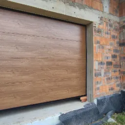 Segmentowa brama garażowa w kolorze drewna, zamontowana w surowym budynku z czerwonej cegły, widoczna w trakcie budowy.