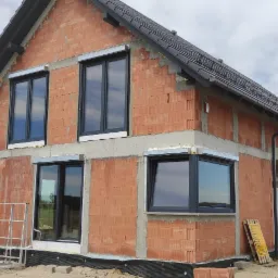 Surowy stan budynku jednorodzinnego z zamontowanymi oknami w grafitowych ramach i częściowo zamontowanymi roletami zewnętrznymi, widoczne pustaki ceramiczne i betonowe nadproża.