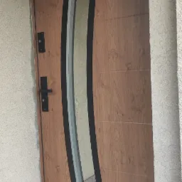 Drewnopodobne drzwi wejściowe z czarnym, łukowatym elementem dekoracyjnym i szybą, zamontowane w ościeżnicy w elewacji o fakturze baranka.