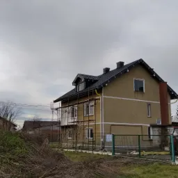 Dwupiętrowy dom z brązową elewacją i nowym, czarnym dachem, częściowo otoczony rusztowaniem, widok z poziomu gruntu w pochmurny dzień.