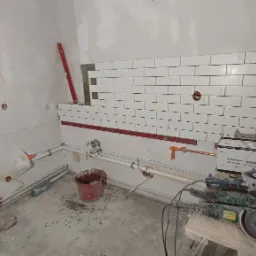 Fragment ściany z częściowo ułożonymi białymi, prostokątnymi płytkami, poziomice w pionie i poziomie, widoczne rury, puszka elektryczna oraz narzędzia do cięcia płytek na prowizorycznym stole.