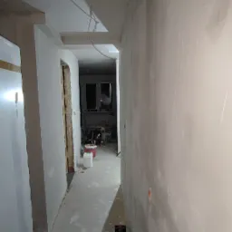 Korytarz w trakcie prac przygotowawczych do malowania, ściany pokryte gładzią, widoczne kable zwisające z sufitu, kubełki z farbą w tle.