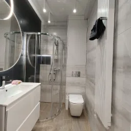 Nowoczesna łazienka z kabiną prysznicową z zaokrąglonymi drzwiami, białą umywalką z szafką, toaletą i grzejnikiem drabinkowym, wykończona płytkami imitującymi marmur i drewnopodobną podłogą.