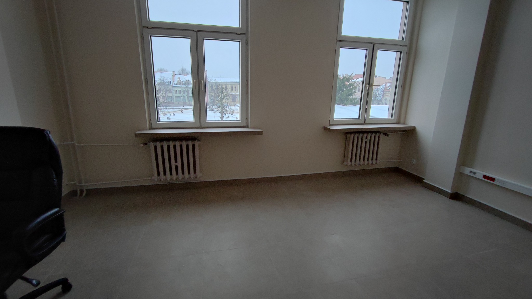 Puste, jasne pomieszczenie biurowe z dwoma oknami, grzejnikami pod oknami i częściowo widocznym czarnym fotelem biurowym po lewej stronie.