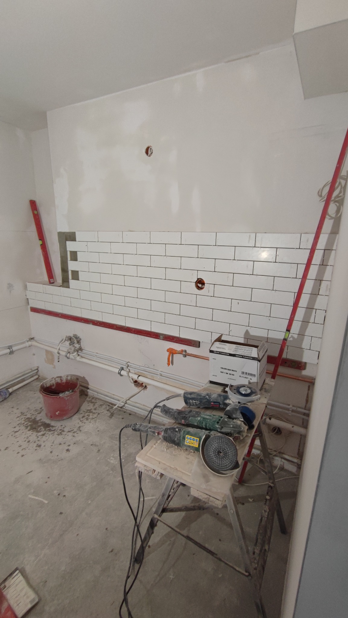Ściana z częściowo ułożoną białą, prostokątną glazurą typu metro, poziomice przylegające do ściany, narzędzia do cięcia glazury na stole warsztatowym, instalacja wodna pod glazurą.