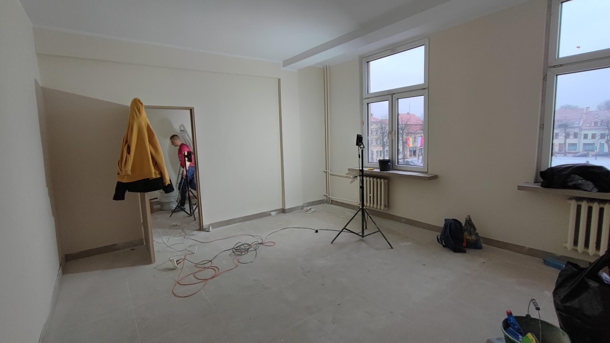 Wnętrze pokoju w trakcie prac, świeżo pomalowane ściany w odcieniach bieli, widoczny pracownik na drabinie, kabel zasilający lampę na trójnogu oraz wiadro z farbą w rogu.