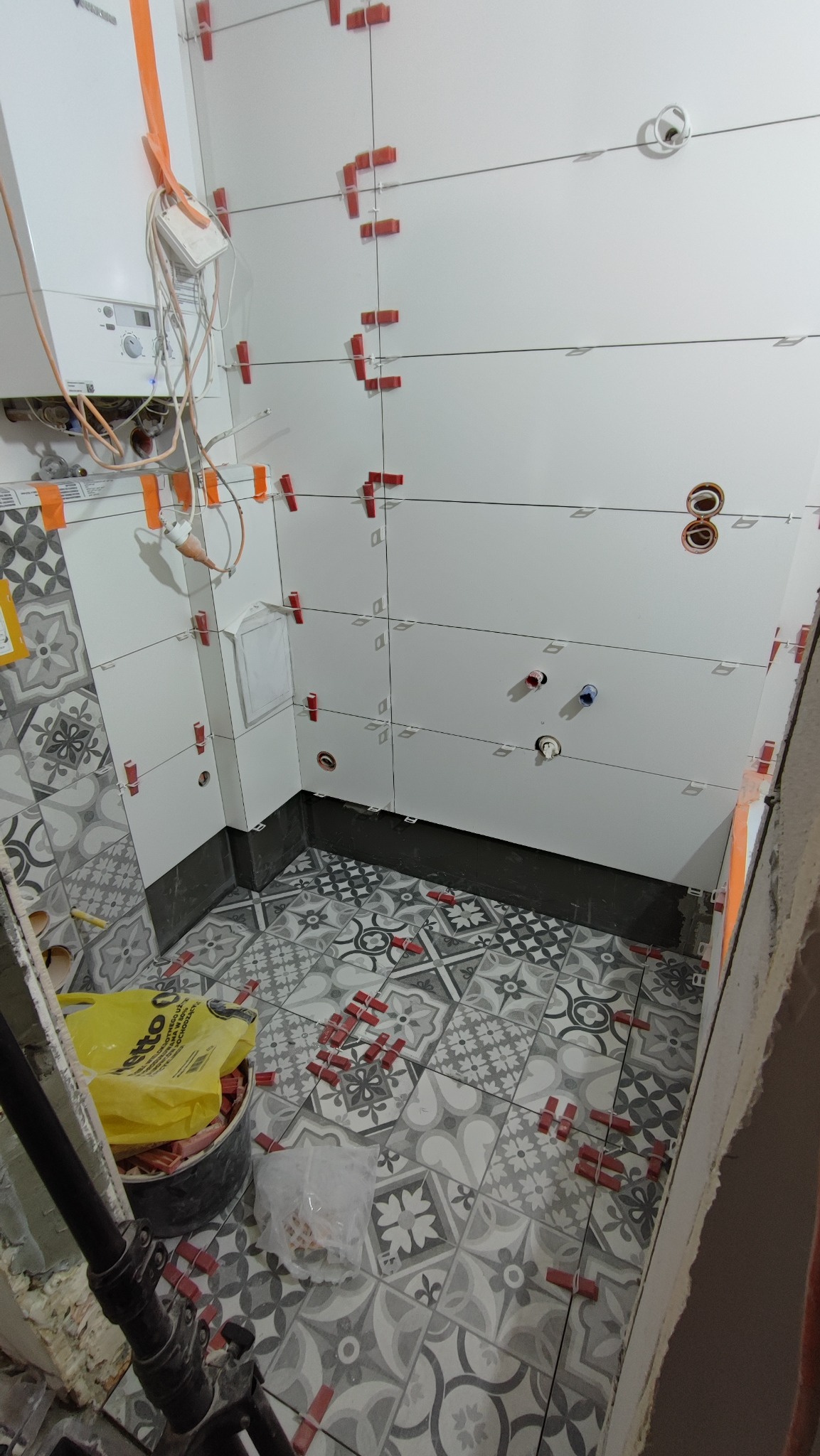 Układanie płytek podłogowych i ściennych w łazience; widoczne białe płytki ścienne z czerwonymi krzyżykami dystansowymi, wzorzyste szaro-białe płytki podłogowe, piec gazowy na ścianie i wiadro...