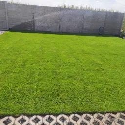 Świeżo założony trawnik z rolki, automatyczne nawadnianie w tle, obrzeża z ażurowych płyt betonowych, szary betonowy płot.