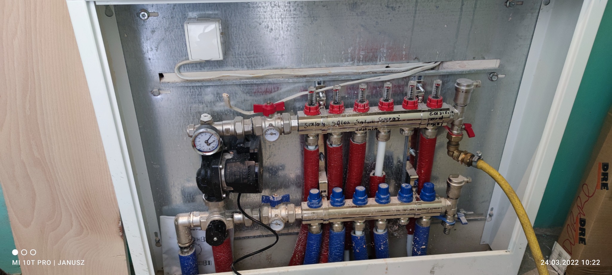 Rozdzielacz instalacji ogrzewania podłogowego w metalowej szafce, widoczne rury w kolorach czerwonym i niebieskim, manometry, zawory oraz pompa obiegowa. Na górnej belce rozdzielacza widoczny...