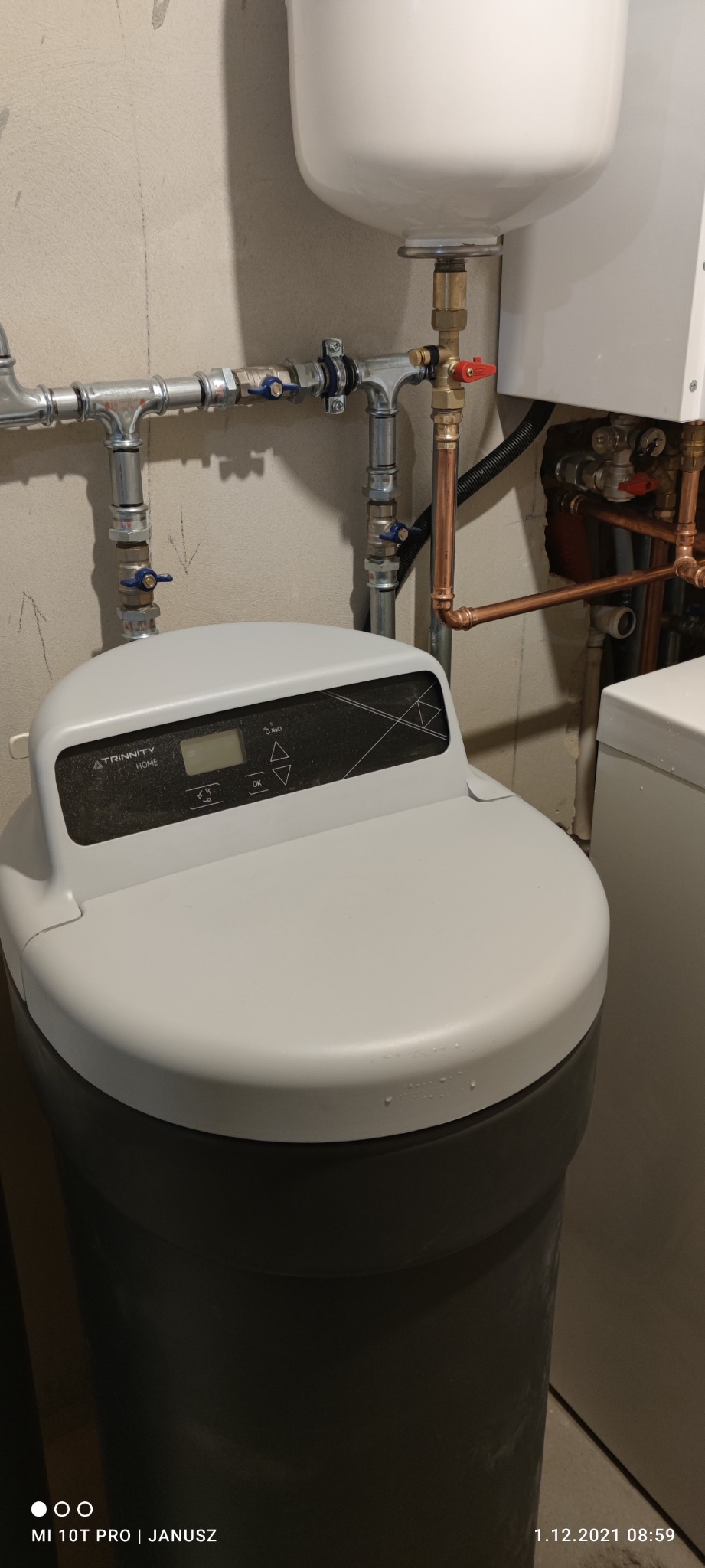 Szary zmiękczacz wody Trinity Home z wyświetlaczem LCD i przyciskami, podłączony do instalacji wodnej z rurami miedzianymi i zaworami, na tle szarej ściany.