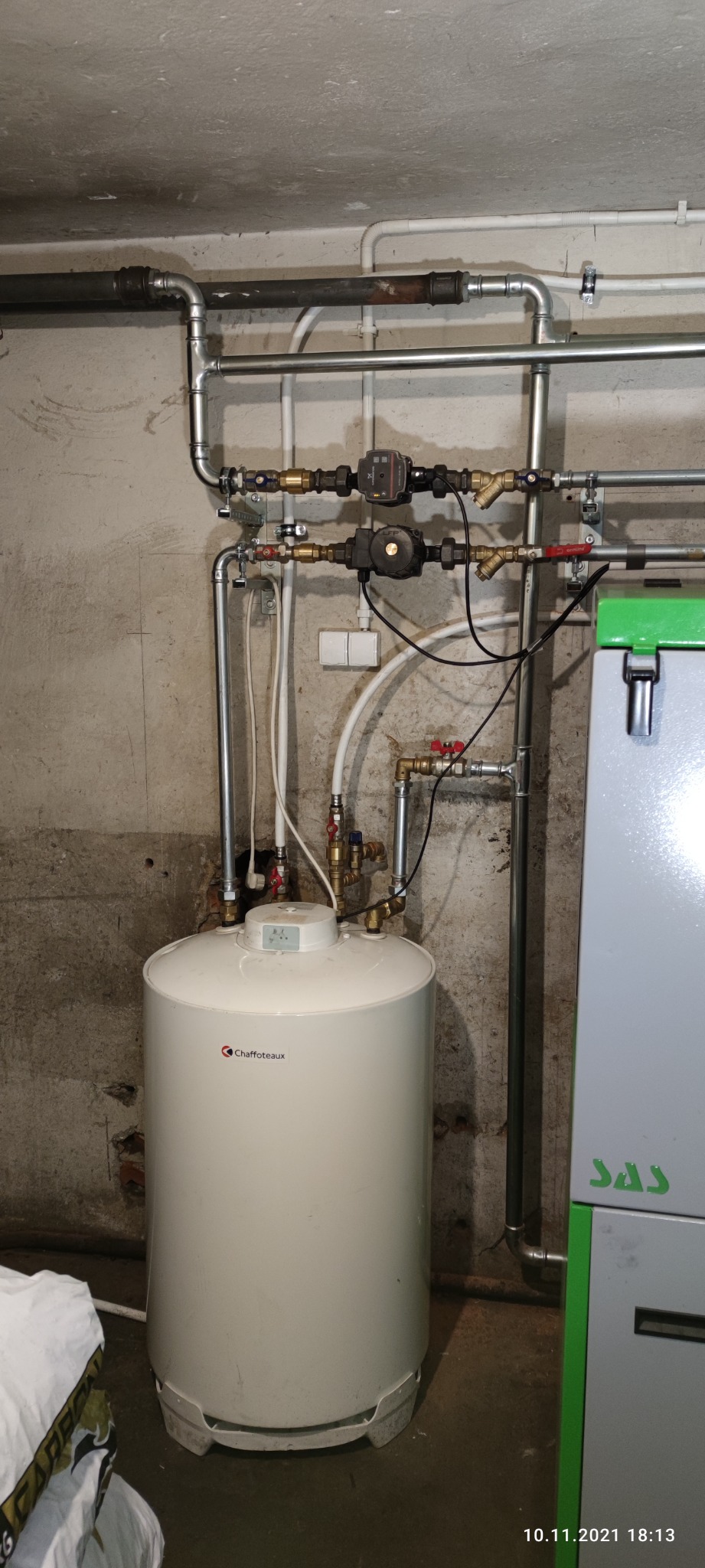 Bojler Chaffoteaux z podłączonymi rurami miedzianymi i zaworami w piwnicy z surowymi ścianami, widoczne pompy obiegowe i fragment pieca SAS.